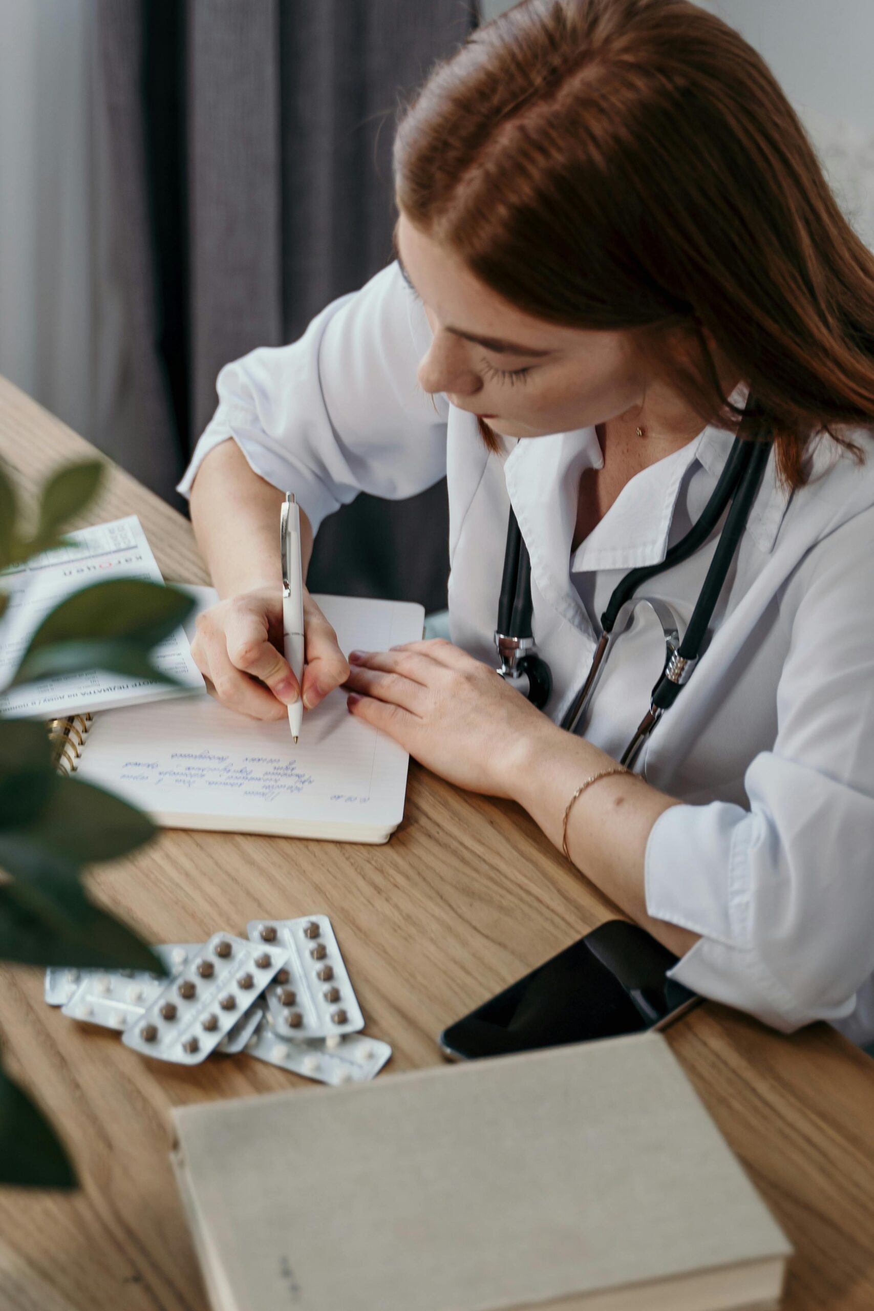 Young female doctor writing a prescription in a medical office with a stethoscope and pills nearby.